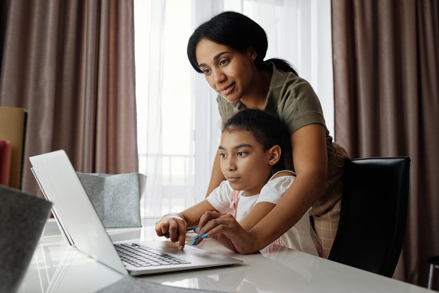 Woman leaning over her child to click on the laptop open on the table as they browse online tutors.