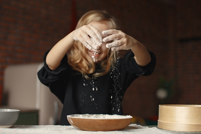 A girl smiling as she lifts her hands with flour from a bowl. She is cooking a recipe from a family-friendly blog.