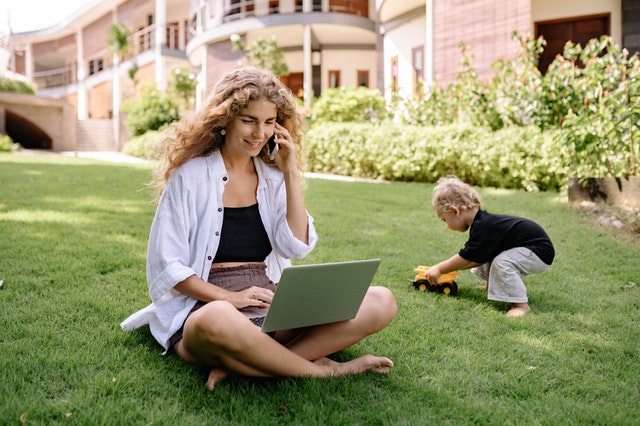 A woman outside in the grass with a child, phone and laptop talking using a US virtual phone system provider.